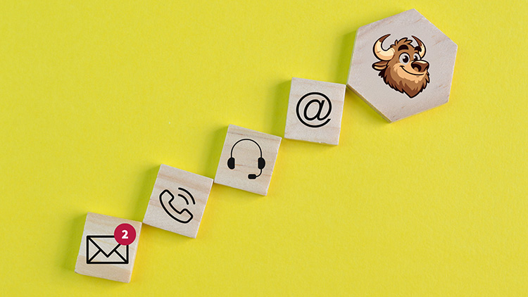 Image showing wooden tiles arranged in a diagonal line on a yellow background. The tiles display icons representing communication methods: an envelope with a notification badge, a phone, a headset, and an email '@' symbol. At the end of the line is a hexagonal tile featuring the Buffaly logo with a cartoon buffalo head, symbolizing Buffaly's customer support or communication features.
